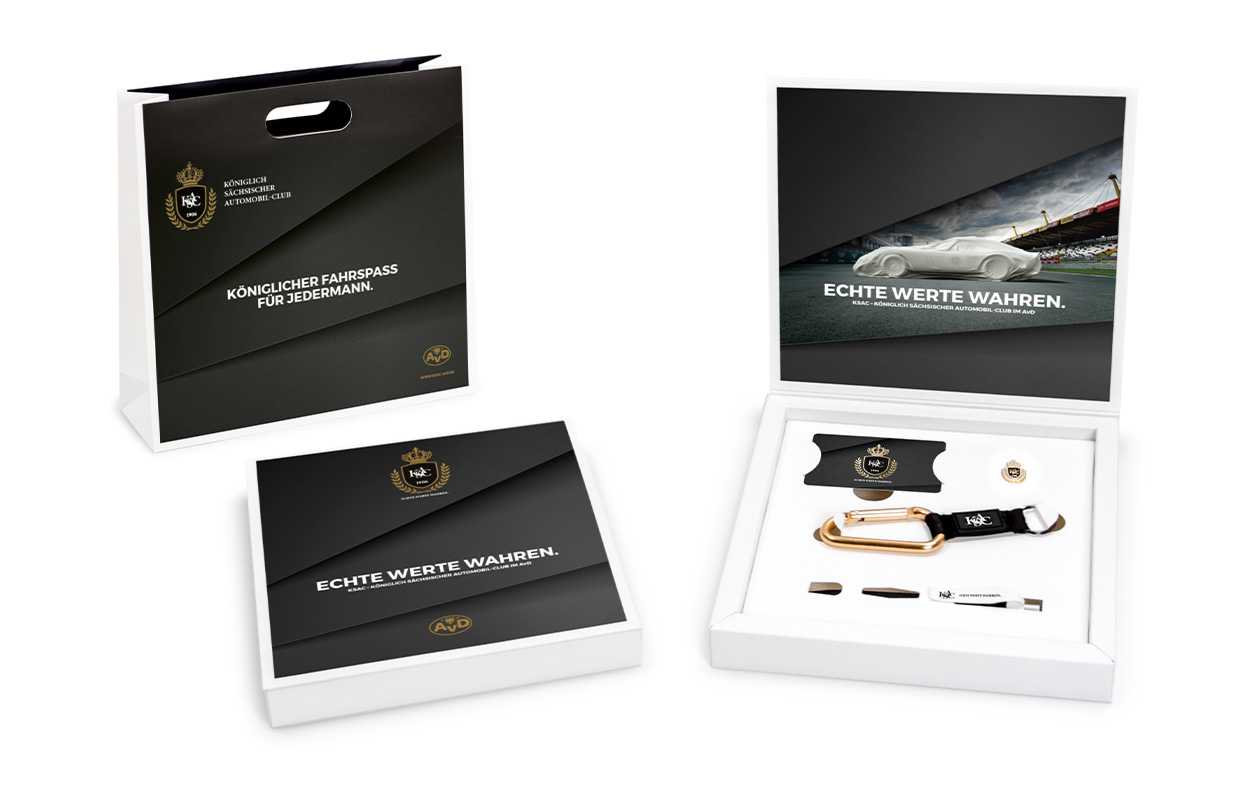 Ein Vollblut KSAC-Geschenkset mit Markenlogo enthält eine Papiertüte und eine offene Schachtel mit Schlüsselanhänger, USB-Stick, Anstecknadel und Lederanhänger. Die Verpackung ist schwarz mit goldenen Akzenten und deutschem Text gestaltet und unterstreicht die AvD-Partnerschaft.