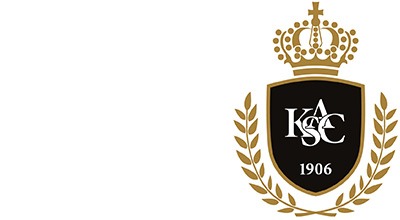 Ein schwarz-goldenes Wappen mit einer stilisierten Krone, Lorbeerzweigen und den Buchstaben „K.A.S.E.“ mit der Aufschrift „1906“ darunter symbolisiert das Emblem eines Fußballvereins. Akzente von Vollblut KSAC AvD bereichern das Design. Weißer Hintergrund.